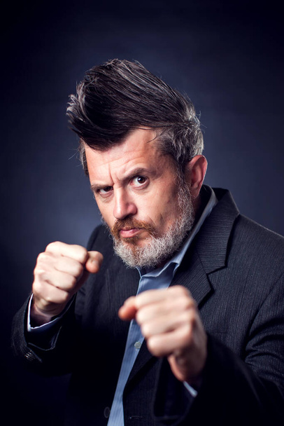 A portrait of bearded man with iroquois wearing suit showing fist gesture in front of black background. People and emotions concept - Photo, Image