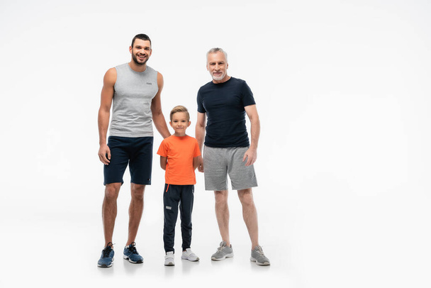 šťastný chlapec s tátou a dědečkem ve sportovním oblečení při pohledu na kameru na bílém - Fotografie, Obrázek