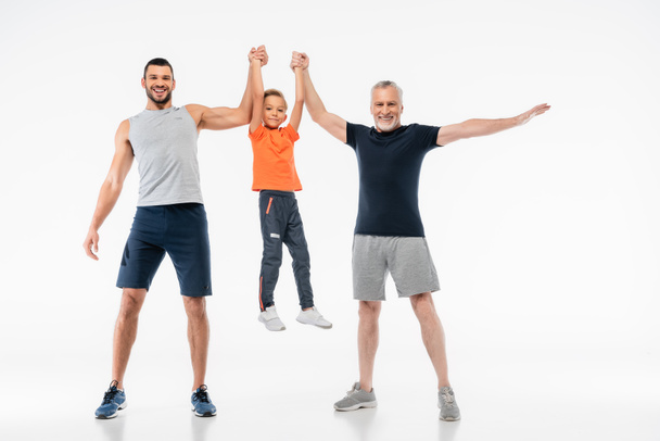 cheerful father and granddad in sportswear holding boy and smiling at camera on white - Photo, Image
