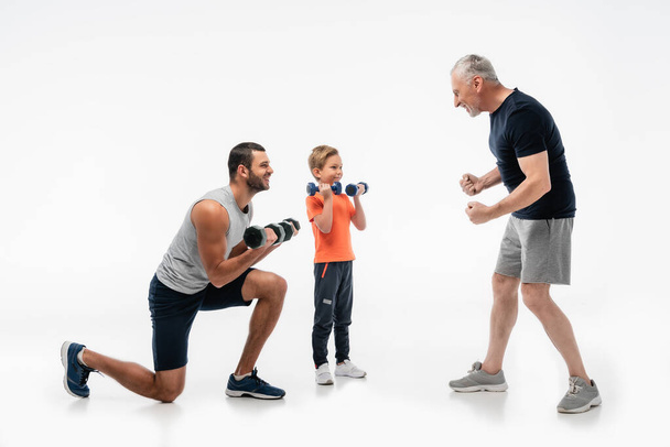 happy man demonstrating strength while father and son exercising with dumbbells on white - Φωτογραφία, εικόνα