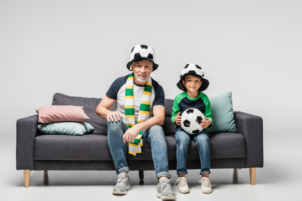 smiling grandfather and grandson watching football match while sitting on sofa in fan hats on grey - Φωτογραφία, εικόνα