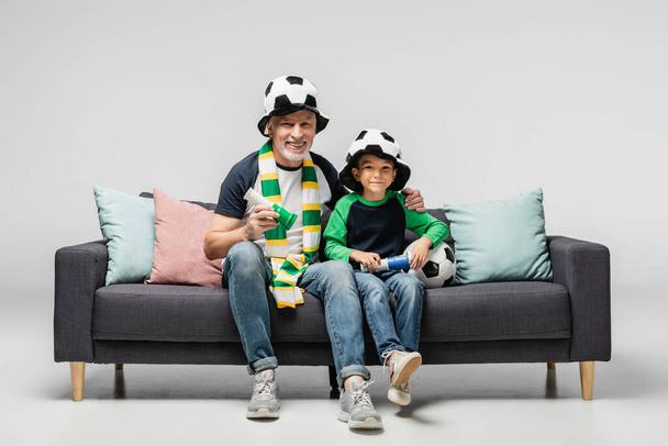 happy man and grandson in football fan hats holding horns while looking soccer match on grey - Фото, зображення