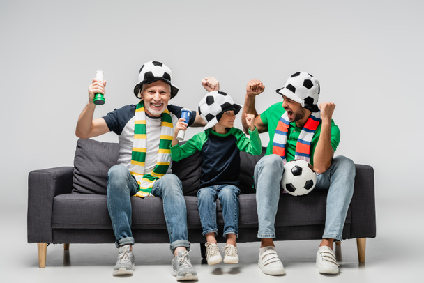 excité garçon avec grand-père et papa montrant geste de victoire tout en regardant match de football dans les chapeaux de ventilateur sur gris - Photo, image