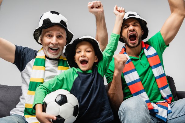 šťastný muži a chlapec v klobouky fanoušků křičí a ukazuje vítězství gesto při sledování fotbalového utkání izolované na šedé - Fotografie, Obrázek