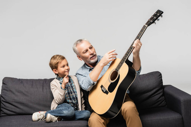 dojrzały mężczyzna wskazując palcem na gitarę akustyczną w pobliżu wnuka odizolowany na szarości - Zdjęcie, obraz