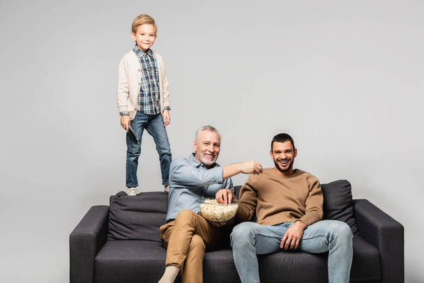 smiling boy standing on sofa near dad and grandfather watching tv and eating popcorn isolated on grey - Zdjęcie, obraz