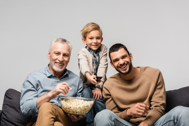 cheerful boy holding remote controller near happy grandfather and dad watching tv with popcorn isolated on grey - Фото, зображення