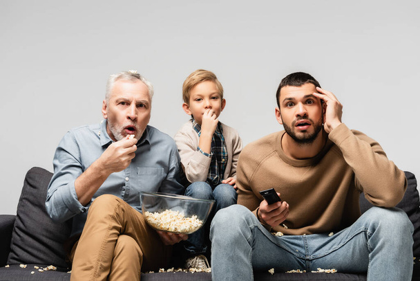 hämmästynyt mies pitää kulhoon popcornia katsellessaan televisiota yhdessä poika ja pojanpoika eristetty harmaa - Valokuva, kuva