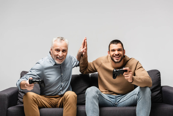KYIV, UKRAINE - NOVEMBER 17, 2020: excited father and son giving high five while sitting on sofa with joysticks isolated on grey - Fotografie, Obrázek