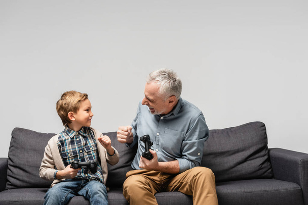 KYIV, UKRAINE - NOVEMBER 17, 2020: happy grandfather and grandson doing fist bump while holding joysticks isolated on grey - Foto, Imagen