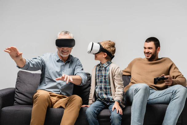 KYIV, UKRAINE - NOVEMBER 17, 2020: mature man gesturing in vr headset near grandson and son with joystick isolated on grey - Photo, Image