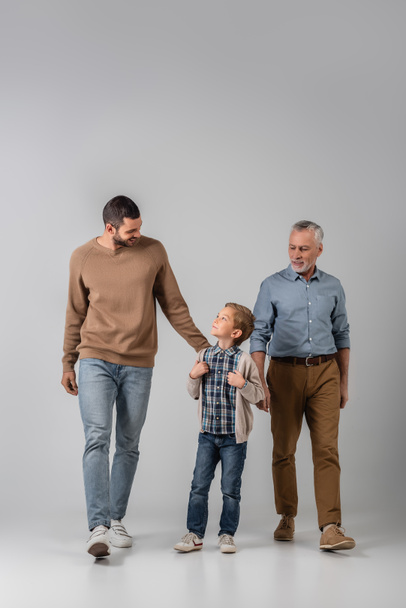 happy boy looking at father while walking near grandpa on grey - Foto, Bild