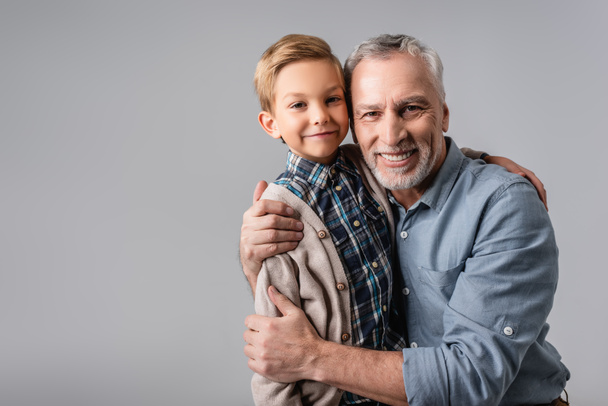 feliz hombre maduro abrazando nieto sonriente mientras mira a la cámara aislada en gris - Foto, imagen