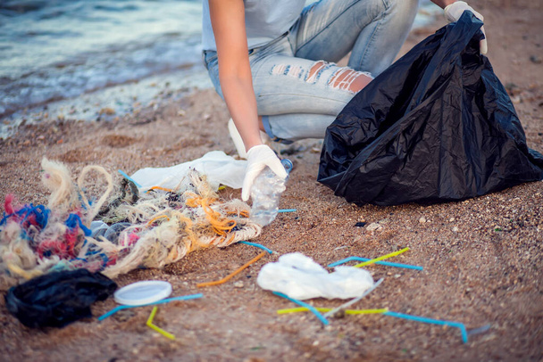 Жінка в синій сорочці з білими рукавичками і великим чорним пакетом, що збирає сміття на пляжі. Концепція захисту навколишнього середовища та забруднення планети
 - Фото, зображення