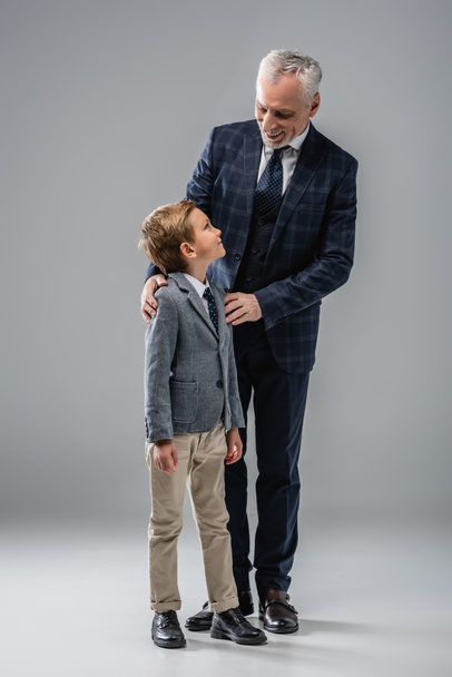 sonriente hombre de negocios en formal desgaste abrazando hombros de nieto en gris - Foto, imagen