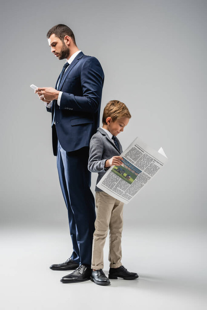 businessman messaging on smartphone while standing back to back with son reading newspaper on grey - Foto, imagen