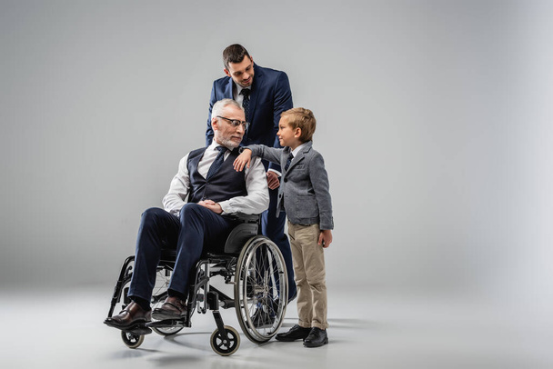 χαμογελαστός άνδρας με επίσημη ένδυση στέκεται κοντά στο γιο και τον πατέρα σε αναπηρική καρέκλα σε γκρι - Φωτογραφία, εικόνα