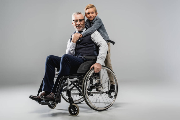 χαμογελαστό αγόρι αγκαλιάζει παππού σε αναπηρική καρέκλα, ενώ κοιτάζοντας κάμερα μαζί σε γκρι - Φωτογραφία, εικόνα
