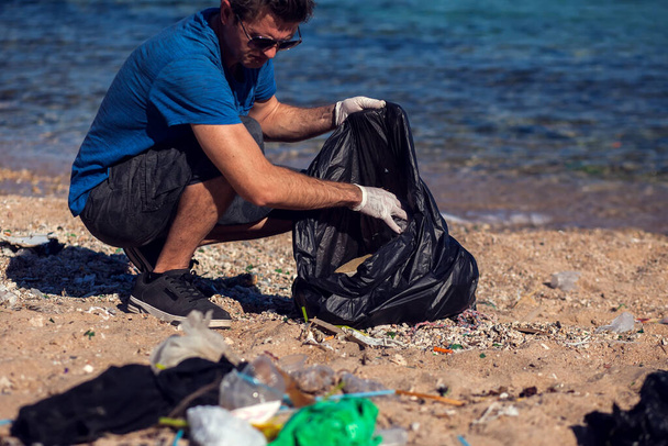Siyah çantalı gönüllü bir adam sahilde çöp topluyor. Çevre kirliliği kavramı - Fotoğraf, Görsel