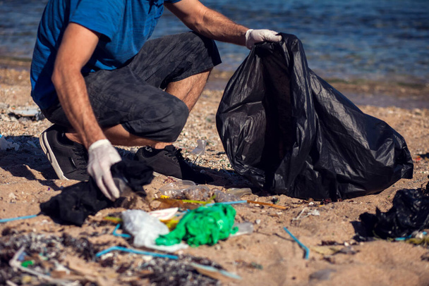 Siyah çantalı gönüllü bir adam sahilde çöp topluyor. Çevre kirliliği kavramı - Fotoğraf, Görsel