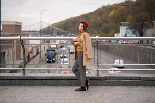 Okouzlující francouzská mladá žena pije kávu pomocí hrnku s kávou při pohledu na kameru usmívající se na pěší most s auty na silnici. Hledáte šťastný módní dívka v červeném baretu a béžový kabát - Fotografie, Obrázek