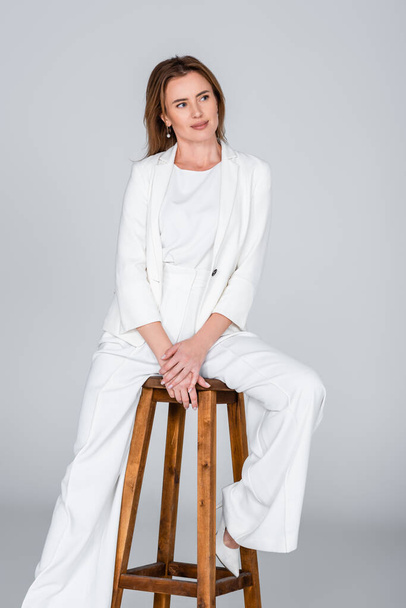 Nainen valkoisessa asussa istuu jakkaralla ja katselee pois eristettynä harmaalla - Valokuva, kuva