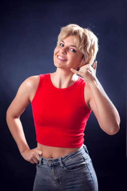 Un portrait de jeune femme aux cheveux blonds courts en chemise rouge montrant parler par téléphone symbole devant fond noir. Style de vie, émotions et concept de communication - Photo, image