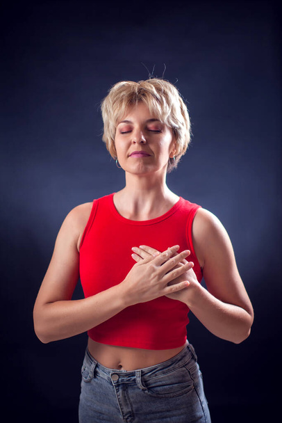 A portrait of young woman with short blond hair in red shirt keeps hands on heart in front of black background. People, emotions and beauty concept - Foto, Imagen
