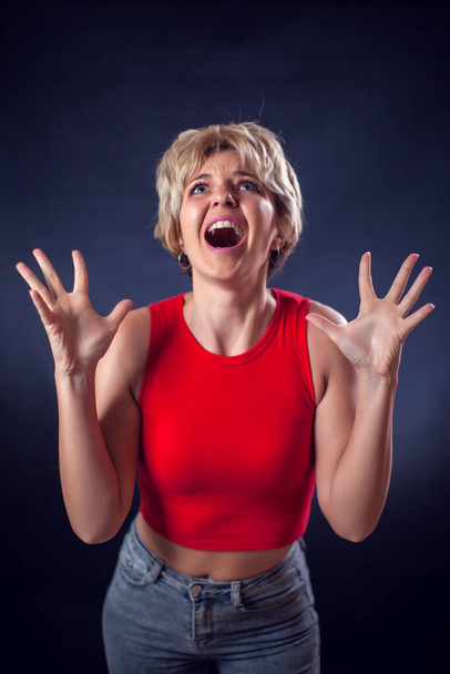 A portrait of screaming young woman with short blond hair in red shirt in front of black background. People and emotions concept - Foto, afbeelding