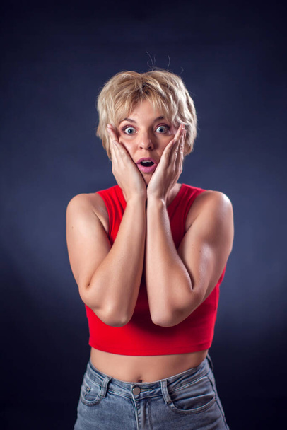 A portrait of young woman with short blond hair in red shirt feeling panic in front of black background. People, emotions and beauty concept - Foto, Bild