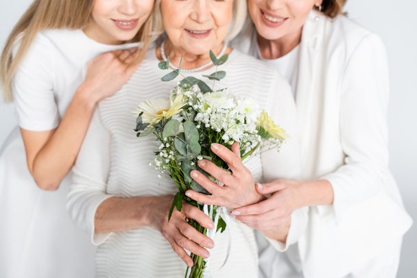 abgeschnittene Ansicht einer zufriedenen Seniorin, die lächelt, während sie Blumen in der Nähe von Tochter und Enkelin hält, isoliert auf Weiß - Foto, Bild