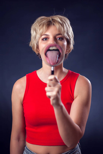 A portrait of young woman with short blond hair in red shirt holding magnifier in front of opened mouth with tongue beside of black background. Lifestyle and emotions concept - Фото, зображення