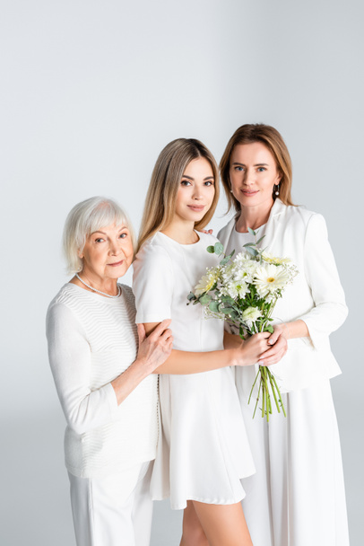 happy young woman smiling while holding flowers near mother and granny isolated on grey - Photo, Image