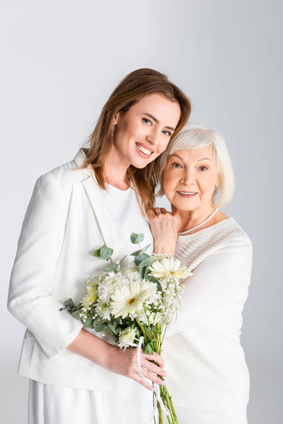 cheerful daughter holding flowers and smiling with happy senior mother isolated on grey - Photo, Image