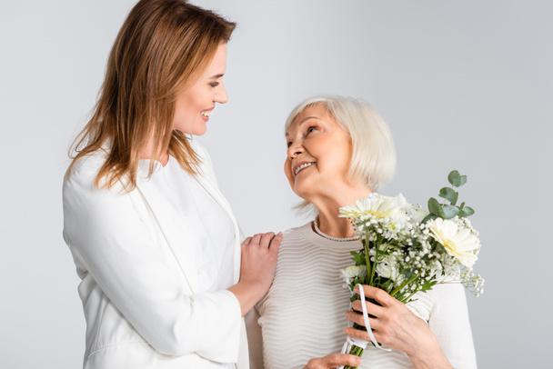 cheerful daughter looking at happy senior mother with flowers isolated on grey - Photo, Image