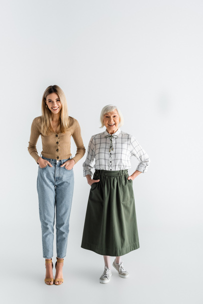 full length of happy granddaughter and granny standing with hands in pockets on white - 写真・画像