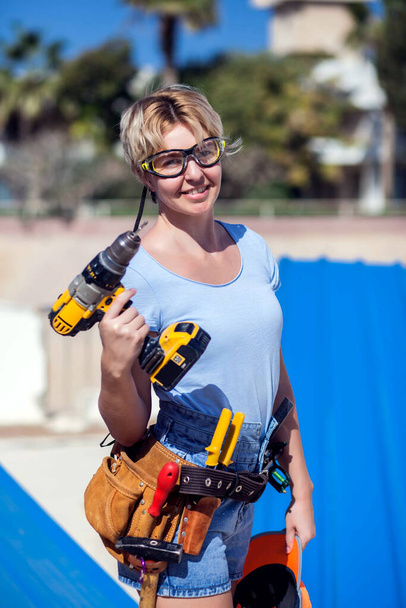 Handige vrouw met gereedschapsriem en gele boor in haar handen buiten - Foto, afbeelding