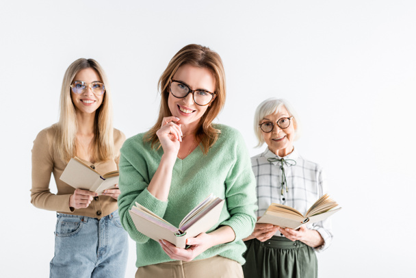 three generation of smiling women in glasses reading books isolated on white - Фото, изображение