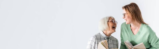 senior mother laughing with daughter in glasses while holding books isolated on white, banner - Foto, Imagem