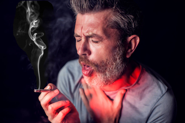 Mies polttaa tupakkaa ja hänellä on ongelmia keuhkojen kanssa mustan taustan edessä. Tupakkariippuvuuden käsite - Valokuva, kuva