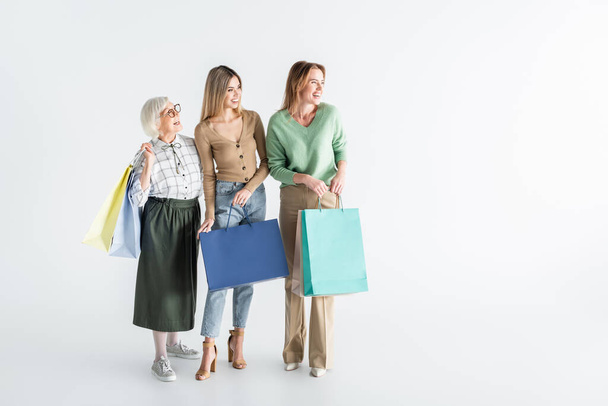 full length of three generation of happy women holding shopping bags and looking away on white - Foto, Imagen