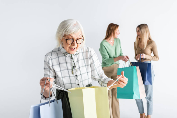 surprised senior woman looking at shopping bag near daughter and granddaughter on blurred background - Photo, Image