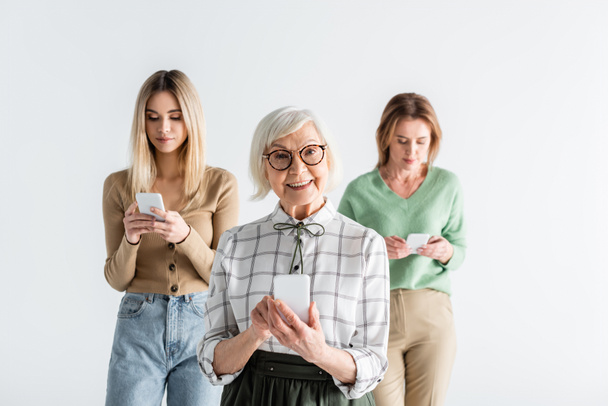 cheerful senior woman in glasses holding smartphone near daughter and granddaughter on blurred background - Fotografie, Obrázek