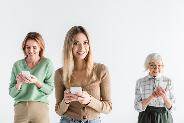 šťastná mladá žena stojící s chytrým telefonem v blízkosti matky a babičky na rozmazaném pozadí izolované na bílém - Fotografie, Obrázek