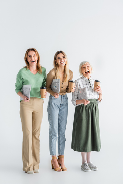full length of three generation of cheerful women holding newspapers and paper cups on white - Fotó, kép