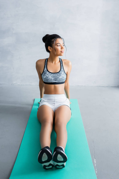 young african american woman in sportswear looking away while sitting on fitness mat at home - Foto, Bild