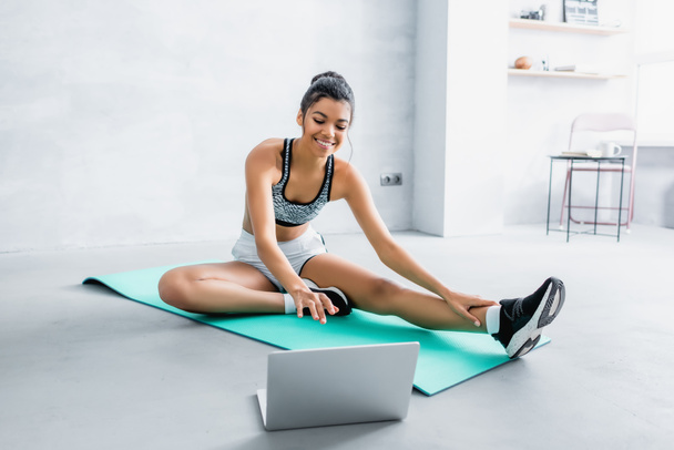 sonriente mujer afroamericana en ropa deportiva sentado en la alfombra de fitness cerca de la computadora portátil en casa - Foto, imagen