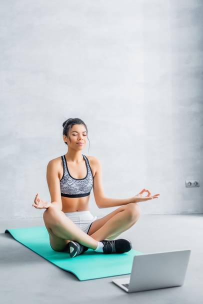 young african american woman in sportswear meditating in lotus pose on fitness mat near laptop - Photo, Image