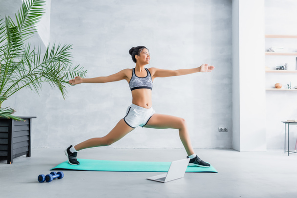 cheerful african american woman in sportswear practicing warrior pose near laptop at home - Foto, imagen