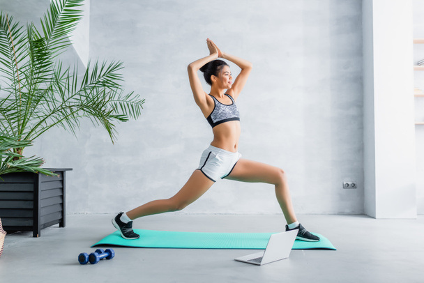 young african american sportswoman exercising in warrior pose near laptop and dumbbells at home - Foto, afbeelding